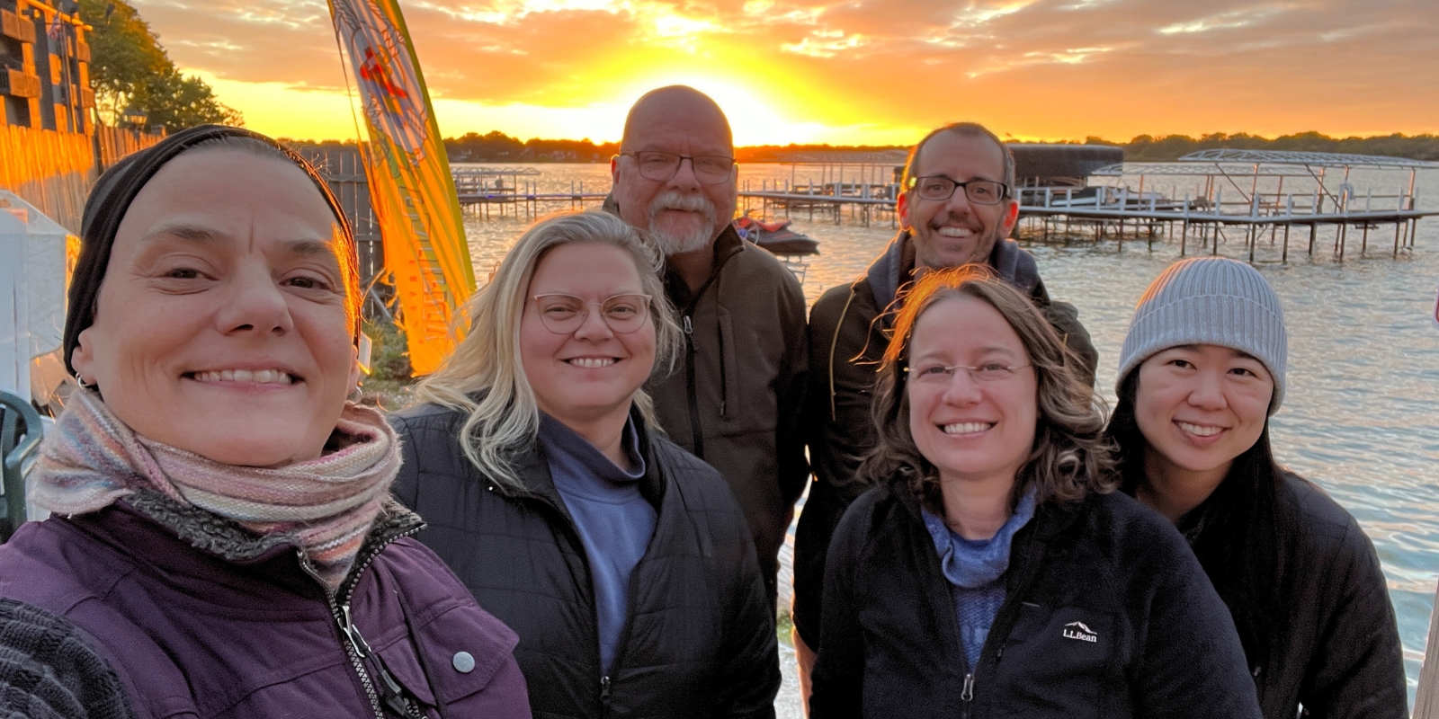 AOS Staff at 2023 Retreat in Illinois. From left to right: Chris Anne, Crystal, Mark, Chris, Judith, and Mint with sunset over lake in background