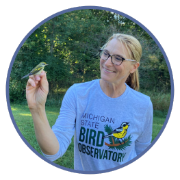 Jennifer Owen smiling at warbler she's holding in her hand while wearing Michigan State Bird Observatory t-shirt